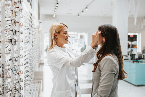 Beautiful and fashionable woman choosing eyeglasses frame in modern optical store. Female seller specialist helps her to make right decision.