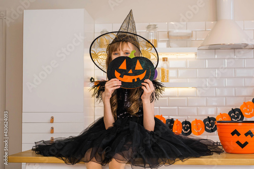 Funny blonde girl kid 8-9 years old in a witch costume sitting on the kitchen table decorated for the Halloween holiday. Traditions, holidays, scenery concept. photo