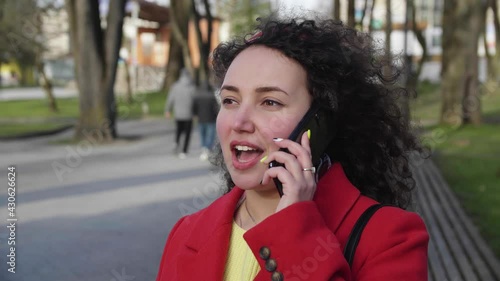 Beautiful young curly girl in a red coat is chatting on a smartphone in the city
