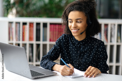 Satisfied friendly smart african american young woman with headphones, office worker or support center operator, sits at her desk, conducts online consultation with client, taking notes, smiles