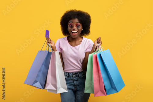 Overjoyed african american lady with shopping bags and credit card smiling over yellow background, studio shot © Prostock-studio