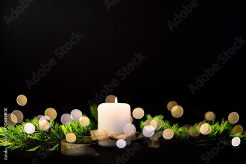 Scena con una candela bianca accesa legata con un fioco dorato e circondata di rami verdi di abete  su uno sfondo nero con spazio per scrivere. Condoglianze. Auguri.