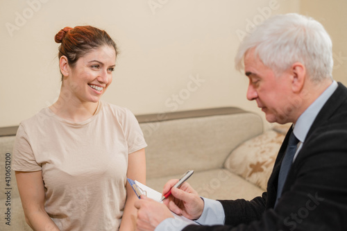 Mature gray-haired male psychotherapist with clipboard in session with female patient. Smiling caucasian woman at a psychologist appointment.