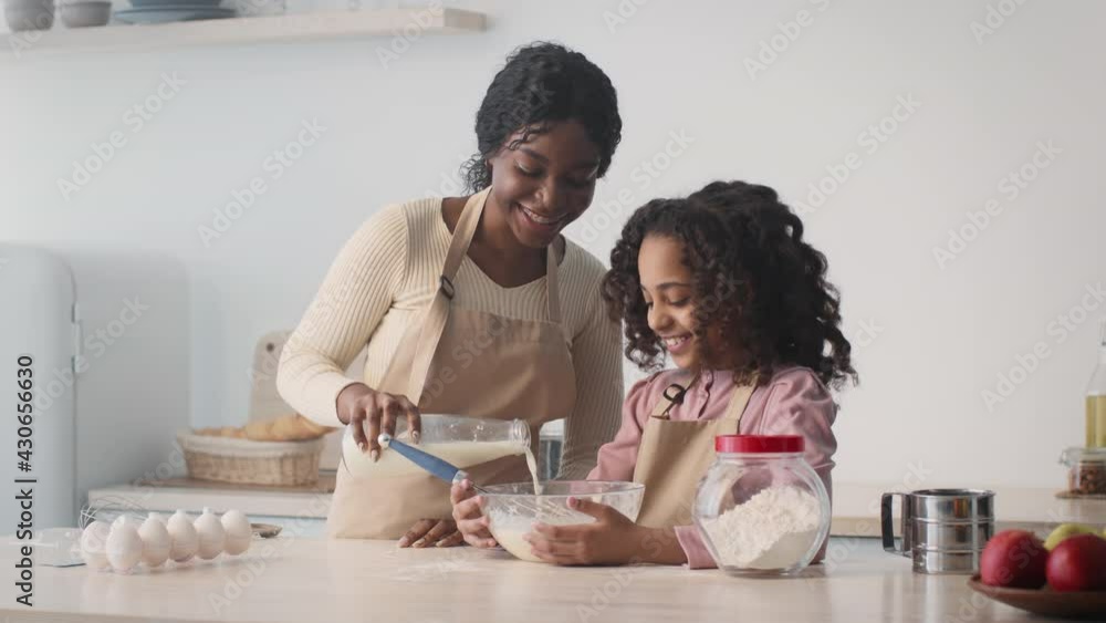 Happy african american mother and daughter preparing dough, woman adding milk, girl whisking it in bowl at kitchen Happy african american mother and daughter preparing dough, woman adding milk, girl whisking it in bowl at kitchen
