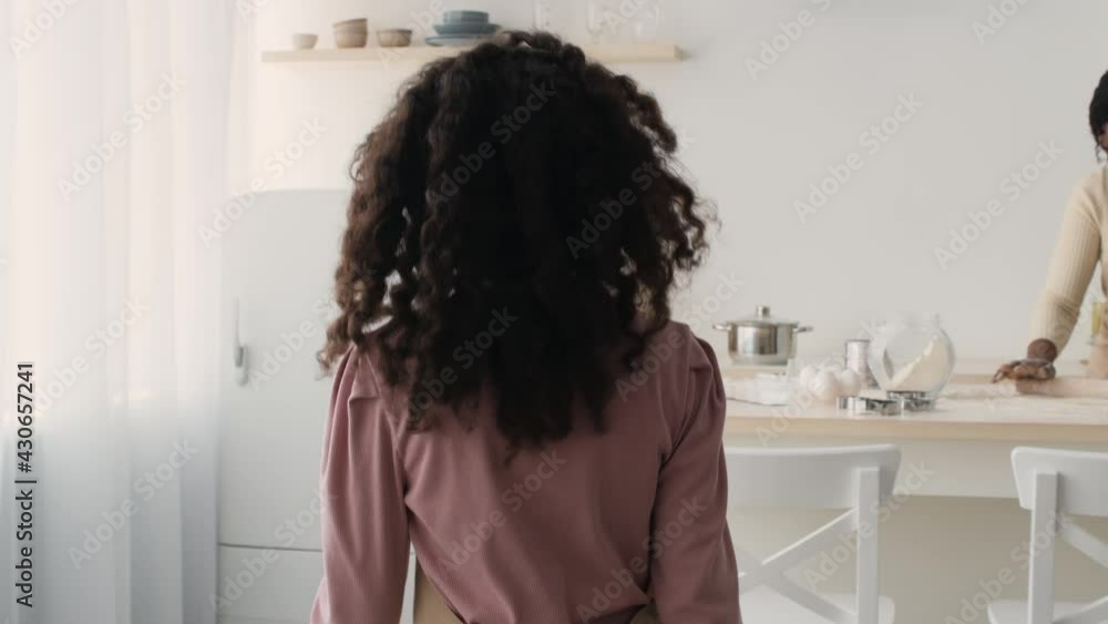 Follow shot of african american girl going to mother and helping her, adding flour to dough at kitchen, slow motion Follow shot of african american girl going to mother and helping her, adding flour to dough at kitchen, slow motion