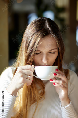 Beautiful attractive business woman in a white blouse drinks cofee in the cafe. Coffee break. Morning light falls from the window