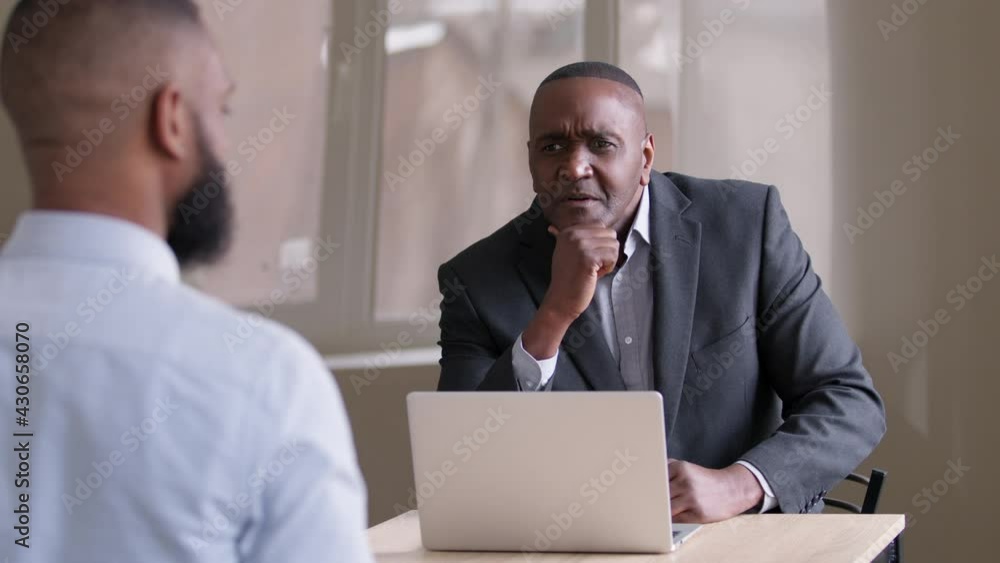 Mature businessman african boss middle age 50s leader sitting at table with laptop talking to unrecognizable man worker at meeting in office at job interview having conversation with unknown afro male Mature businessman african boss middle age 50s leader sitting at table with laptop talking to unrecognizable man worker at meeting in office at job interview having conversation with unknown afro male