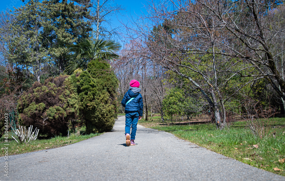 girl in a blue jacket and a pink hat walks along the path in the Arboretum Park on a sunny spring day. Sochi, Russia