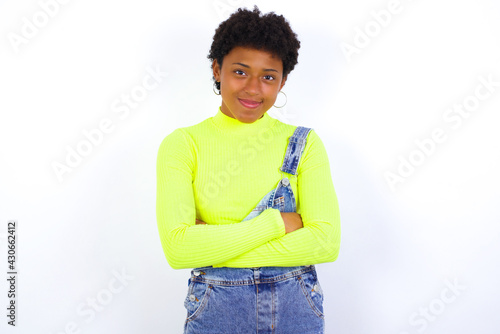 Self confident serious calm young African American woman with short hair wearing denim overall against white wall stands with arms folded. Shows professional vibe stands in assertive pose.
