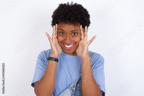 African American female with afro hair wears jeans overalls over white wall Pleasant looking cheerful  Happy reaction