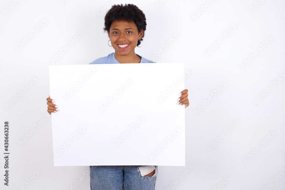 Happy African American female with afro hair wears jeans overalls over white wall Holding Empty Paper Board Advertising Offer Text Standing. Autumn Advertisement Banner. Happy African American female with afro hair wears jeans overalls over white wall Holding Empty Paper Board Advertising Offer Text Standing. Autumn Advertisement Banner.