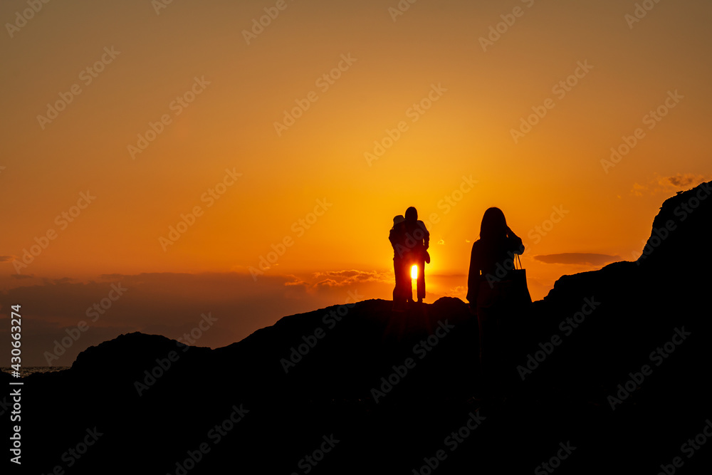 海に沈む夕日を背にインスタ映えする写真を撮影して楽しむ3人の若い女性のシルエット　秋谷海岸　立石公園　梵天の鼻