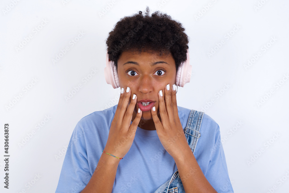 Shocked young African American woman with short hair wearing denim overall against white wall stares fearful at camera keeps mouth widely opened wears wireless stereo headphones on ears Shocked young African American woman with short hair wearing denim overall against white wall stares fearful at camera keeps mouth widely opened wears wireless stereo headphones on ears