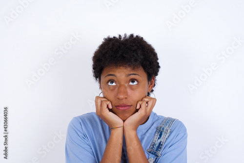 Portrait of sad young African American woman with short hair wearing denim overall against white wall hands face look empty space © Jihan