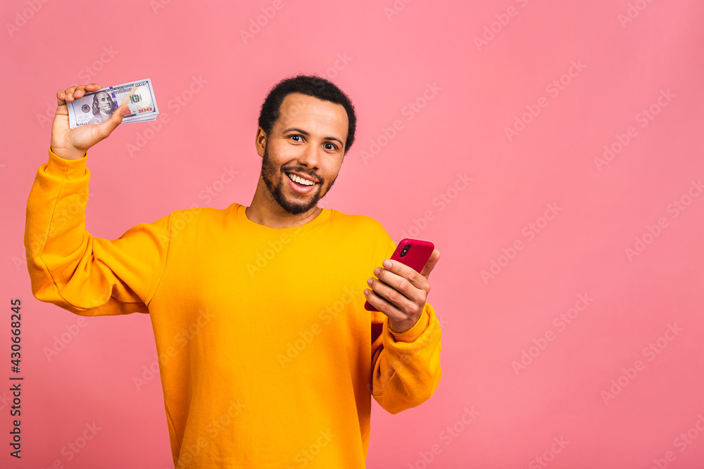 Happy winner! Young rich african american man in casual holding money dollar bills and mobile phone with surprise isolated over pink wall background. Happy winner! Young rich african american man in casual holding money dollar bills and mobile phone with surprise isolated over pink wall background.