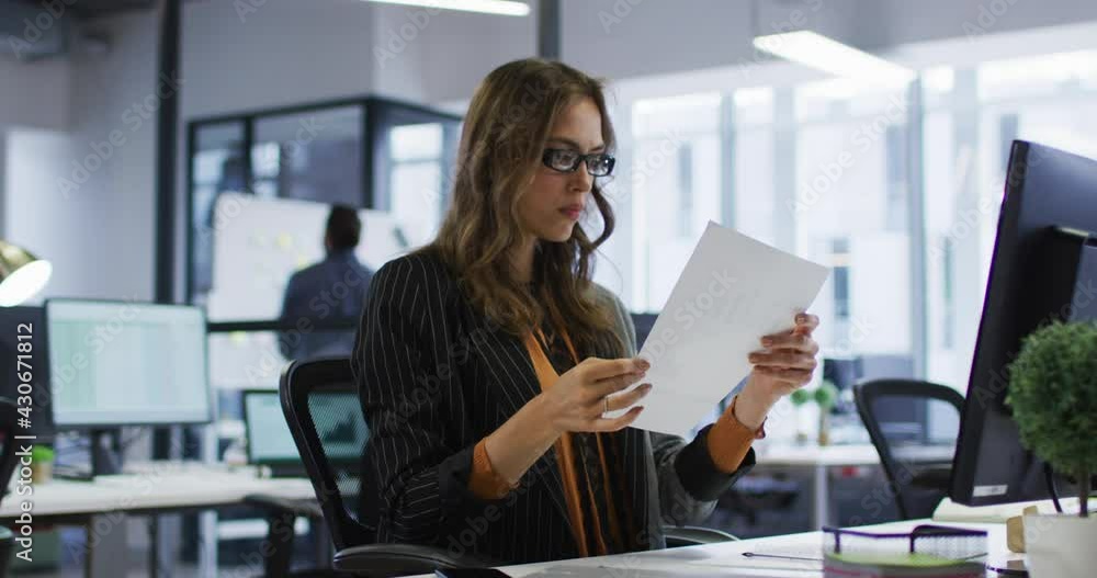 Caucasian businesswoman sitting at desk reading document and taking off glasses in office Caucasian businesswoman sitting at desk reading document and taking off glasses in office