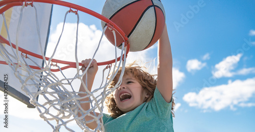 Close up image of kid basketball player making slam dunk during basketball game in floodlight basketball court. The child player is wearing sport clothes.