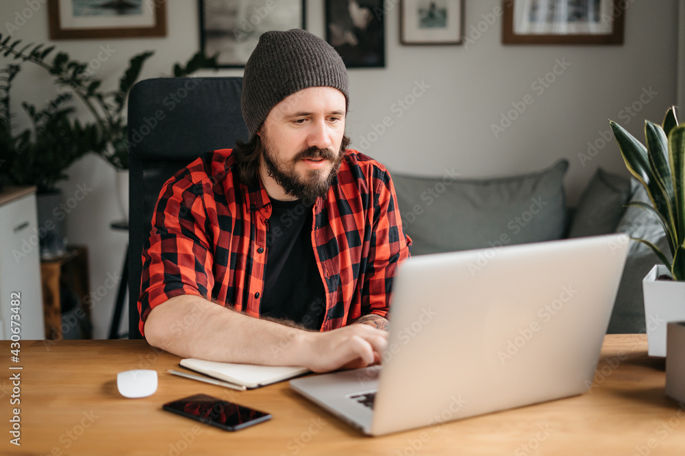 Focused young man with beard dressed in casual clothes using laptop, typing on keyboard, writing email or message, chatting, shopping, successful freelancer working online on computer Focused young man with beard dressed in casual clothes using laptop, typing on keyboard, writing email or message, chatting, shopping, successful freelancer working online on computer