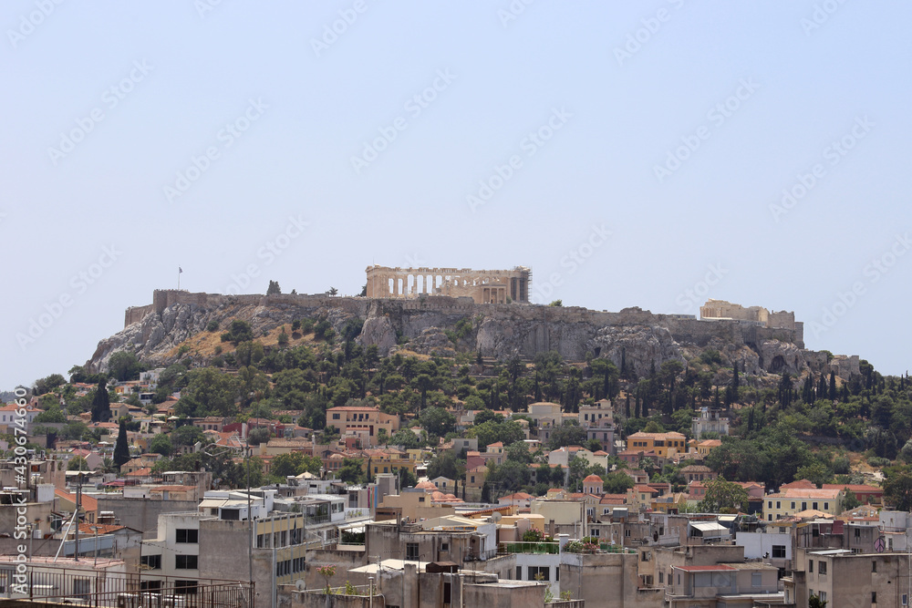 The ancient Acropolis of Athens, Greece and neighborhood below are shown from an elevated view, during daytime in the 2010s. There is room in the blue sky above to add text / copy.