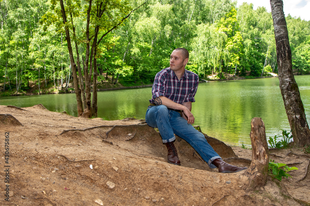 A man in boots and a plaid shirt sat down in the forest by a picturesque pond A man in boots and a plaid shirt sat down in the forest by a picturesque pond