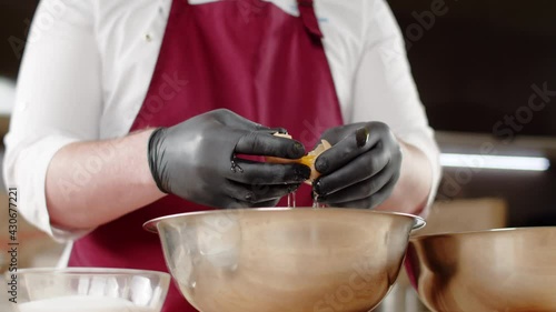 A male chef wearing black gloves breaks an egg and separates the white from the yolk over an iron bowl. A close-up of the separation of the yolk from the protein. Cooking biscuit dough.