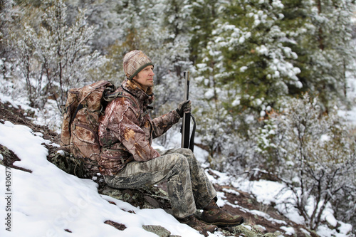 gun hunter wearing camouflage and sitting in snowy scene holding rifle