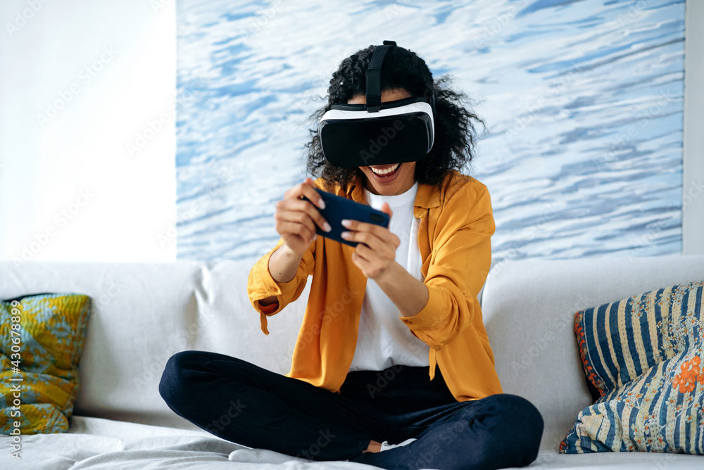 Cyberworld, vr glasses. Joyful cheerful curly haired young african american woman sitting on sofa wearing casual clothes, using smartphone and testing virtual reality device in digital world, smiling
