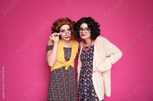 Two cute girl friends woman looking like nerd accountants standing on a pink background. They wear curly brunette wigs and unstylish retro casual outfits. photo