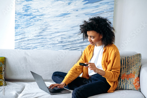 Online shopping concept. Young beautiful african american woman in casual stylish clothes sits on the couch, makes purchases online by laptop, fills in credit card details to pay for goods or delivery photo