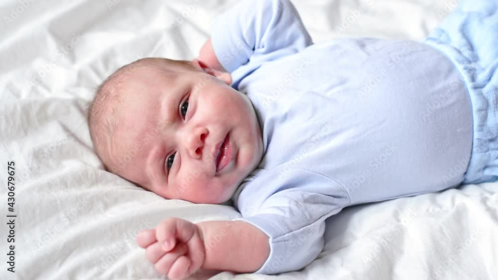 Infant baby waking up. Little child with blue eyes lying on white bed, showing his tongue, and waving his hands. Concept of caring for children and parental love. Portrait of cute baby close-up Infant baby waking up. Little child with blue eyes lying on white bed, showing his tongue, and waving his hands. Concept of caring for children and parental love. Portrait of cute baby close-up