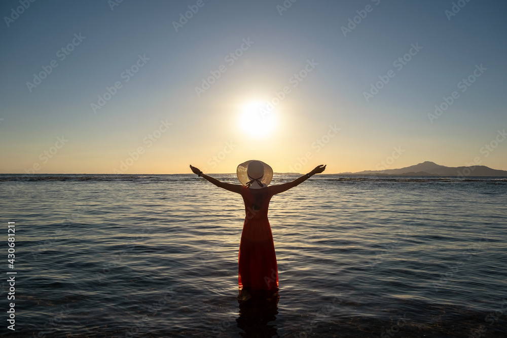 Young woman wearing long red dress and straw hat raising hands standing in sea water at the beach enjoying view of rising sun in early summer morning.