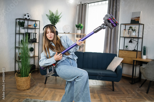 Positive young woman listening music in wireless headphones and playing with handheld vacuum cleaner. Housekeeping with fun. Modern devices concept. photo