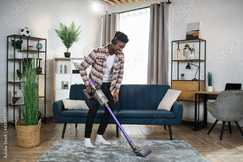 Smiling afro american man in wireless headphones cleaning carpet with handheld vacuum cleaner. Young guy enjoying housekeeping using modern devices. photo