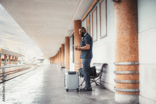A pensive dapper bearded bald African man entrepreneur has missed his train and now watching train schedule on a smartphone while standing on a railway station depot platform with two bags of baggage