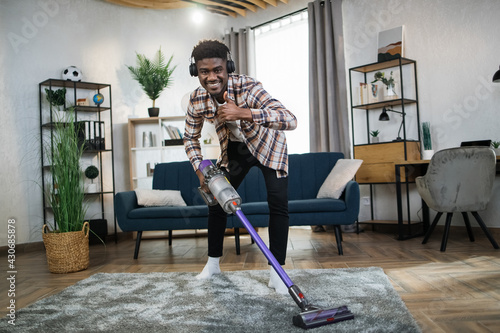 Joyful african guy in headphones smiling and showing thumb up on camera while cleaning apartment with handheld vacuum cleaner. Concept of people, housekeeping and technology.