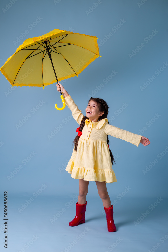 Happy cute little Girl Indian in red rubber boots, cotton dress holds in her hands yellow umbrella zones on a blue background in the studio. concept Spring, space for text - obrazy, fototapety, plakaty 
