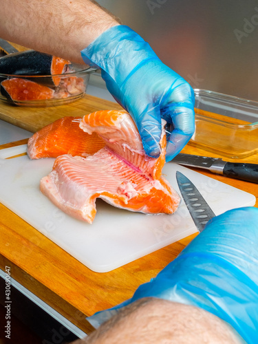 The hands of the cook in hygienic gloves with a sharp knife separate the salmon fillets from the bones. Cutting fresh fish in the kitchen on a cutting board.