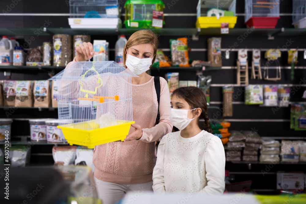 Female customer with small girl in protective face masks buying cage for bird in pet shop