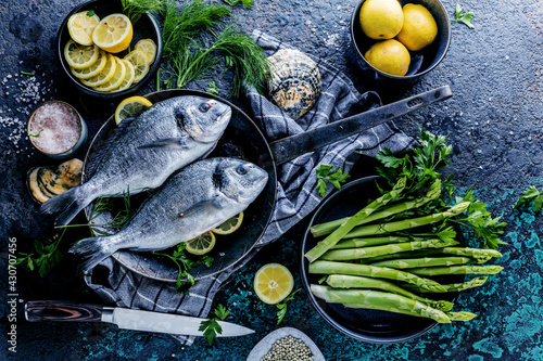 Fresh uncooked dorado or sea bream fish with lemon ,asparagus, herbs, olive oil and spices over black backdrop, top view