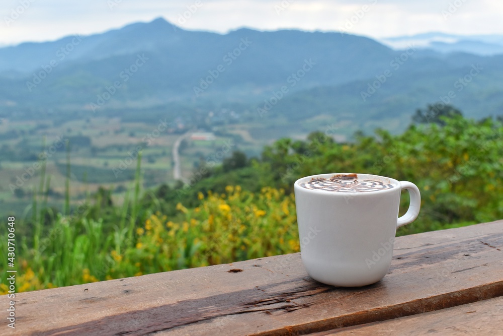 Hot chocolate is placed on a wooden terrace. The background is a mountain and nature view.