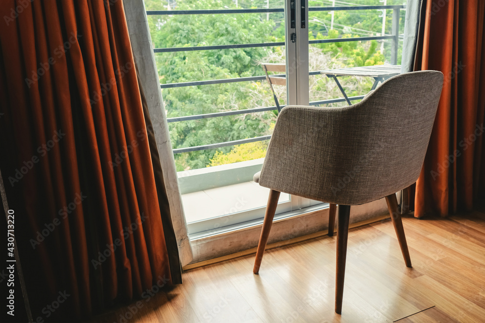 Fototapeta premium Modern chair by the window glass door in the apartment room. The red curtain opened to the morning sunlight. Living in a hotel residence.