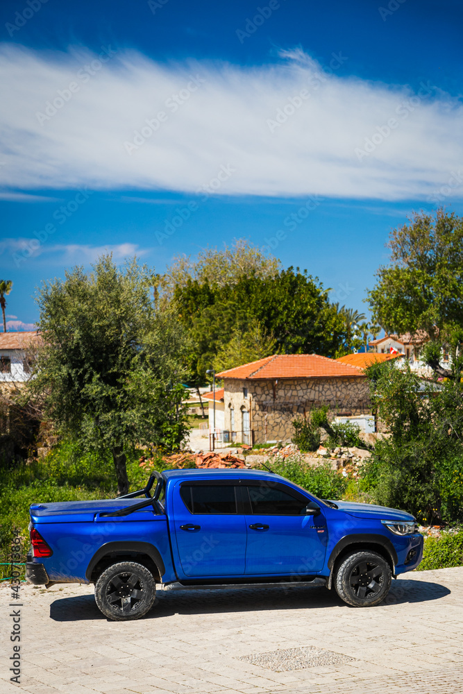 Fototapeta premium Side, Turkey – April 15 2021: blue Toyota Hilux parked on the street on a warm summer day against the backdrop of old street, trees, palms