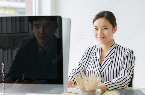 Asian young beautiful female employee wears black and white stripe shirt sit on chair smiling to male worker wears formal suit on opposite office desk who has shadow reflect on computer monitor photo