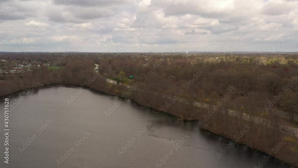 A high angle drone view over a quiet lake, surrounded by dry, brown trees on a cloudy day. The cloudy skies reflect on the water as the camera dolly in and pan left over the water. A high angle drone view over a quiet lake, surrounded by dry, brown trees on a cloudy day. The cloudy skies reflect on the water as the camera dolly in and pan left over the water.