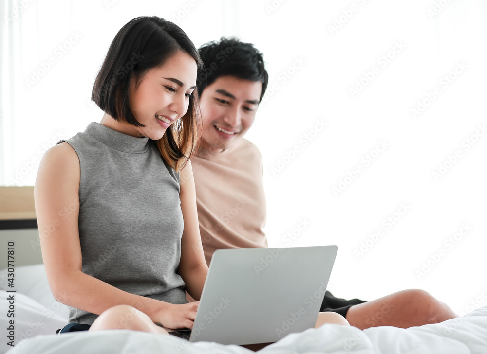 Portrait of cute smiling young Asian lover couple sitting on the bed together. Selective focus on the woman that using digital laptop to doing work at home on weekends. Husband encourages his wife Portrait of cute smiling young Asian lover couple sitting on the bed together. Selective focus on the woman that using digital laptop to doing work at home on weekends. Husband encourages his wife