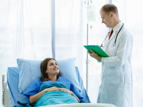 Woman caucasian patient sleep on bed are smiling look happy and comfortable and she look at doctor. Doctor man carrying stethoscope on neck and hold a note to writing about examination