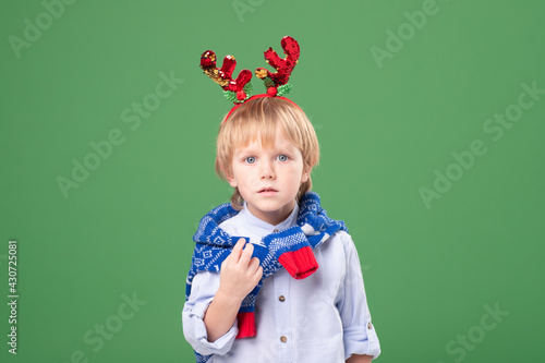 Portrait of a Caucasian blond unhappy child with hair accessory - Christmas deer horns on his head and a winter blue sweater on his neck. Studio green background. Christmas, new year concept. photo