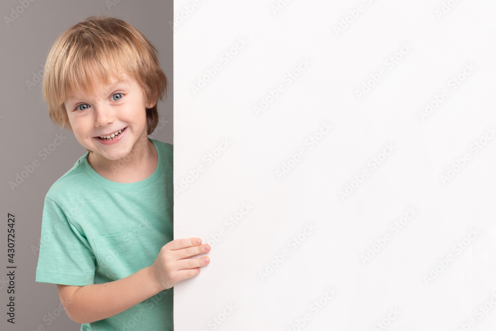 Positive happy caucasian child next to white empty poster. Green T-shirt. Blond hair. Emotional, smiling face. Advertising, educational, dental smile concept. Mock up. Copy space. Positive happy caucasian child next to white empty poster. Green T-shirt. Blond hair. Emotional, smiling face. Advertising, educational, dental smile concept. Mock up. Copy space.