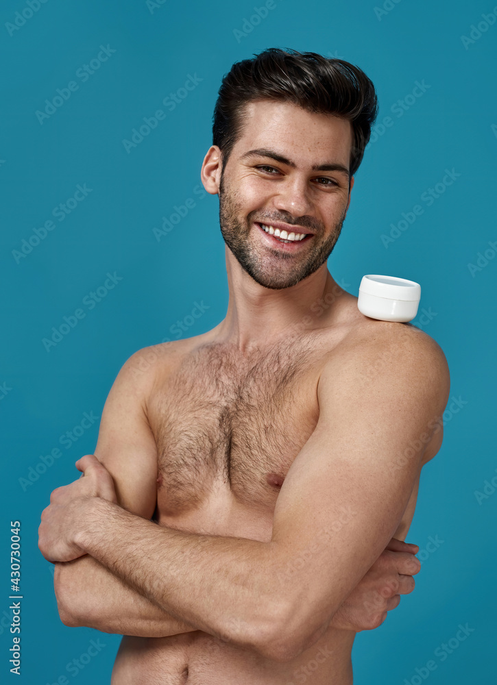 Studio shot of joyful naked man posing with white jar of effective cream on his shoulder, smiling at camera, standing with arms crossed isolated over blue background Studio shot of joyful naked man posing with white jar of effective cream on his shoulder, smiling at camera, standing with arms crossed isolated over blue background