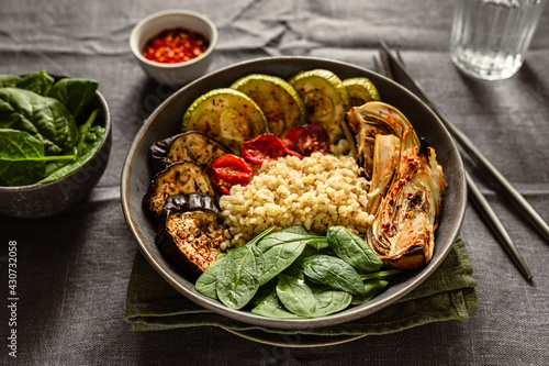 Healthy vegetarian breakfast idea. A bowl with baked vegetables, zucchini, eggplant, tomatoes, fennel, bulgur, fresh spinach. Red hot chili flakes, a glass of water, knife and fork.
